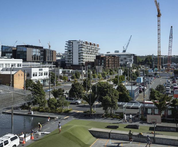 Urban waterfront precinct viewed from an elevated position, with a landscaped recreational area in the foreground containing a basketball half-court and skateboarding features beside the water. Mid-rise apartment and commercial buildings, extensive car parking, roads, mature trees, and several active construction cranes fill the background under a clear sky.
