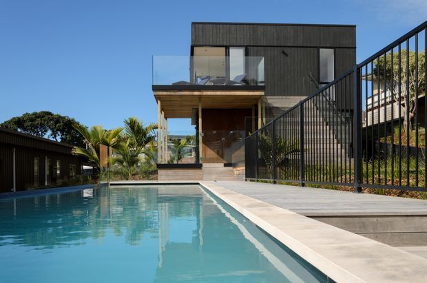 Exterior view of a contemporary multi-level residence beside a swimming pool. The house features dark vertical cladding, extensive glazing, a projecting upper balcony with clear glass balustrades, timber soffits, and black metal fencing. A paved pool deck and landscaped tropical planting frame the scene under a clear blue sky.
