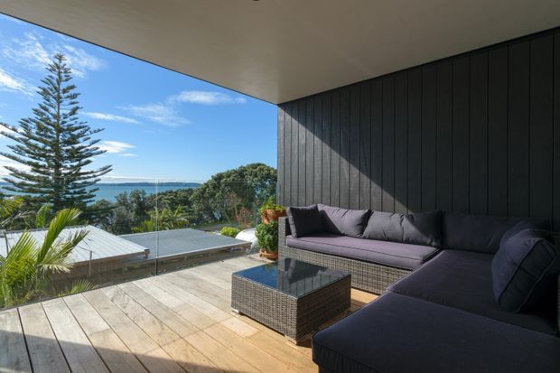 A covered outdoor terrace overlooking a coastal landscape, furnished with a large dark sectional sofa and woven coffee table. The terrace has broad timber decking, a dark vertical-panel feature wall, a smooth light ceiling, and frameless glass balustrading. Bright daylight, blue sky, lush vegetation, and a distant body of water create an open, relaxed setting.