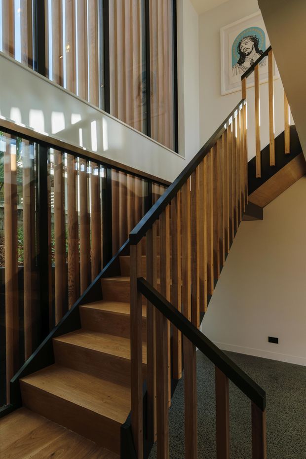 Interior view of a contemporary stairway rising through a bright double-height circulation space. The staircase has warm timber treads and vertical timber balusters, contrasted with dark painted handrails and stringers. Tall glazed openings with vertical timber screening bring in strong sunlight, while pale walls and a grey carpeted landing create a refined, warm atmosphere. Framed artwork is visible on the upper wall.