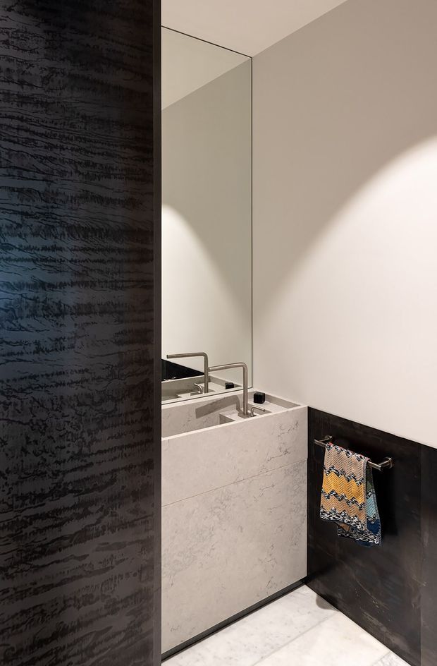 Compact contemporary bathroom with a floating or partially enclosed pale stone vanity, integrated basins, slim brushed-metal tapware, and a large wall mirror. Dark textured wall panels create strong contrast against the light stone surfaces and pale walls, while a patterned hand towel adds a small decorative accent. Dramatic natural light and shadow define the minimalist space.