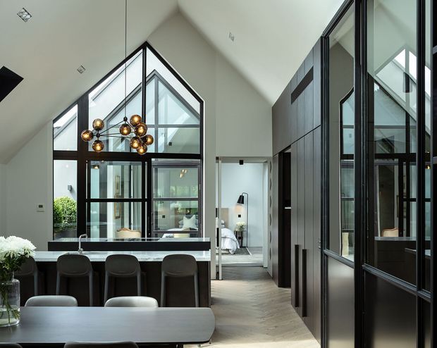 A sophisticated open-plan kitchen and dining space with a dramatic pitched ceiling and extensive black-framed glazing. A dark island with integrated seating sits beneath a sculptural cluster pendant, while a large dining table occupies the foreground. Full-height dark cabinetry and glazed partitions line the right side, and a bedroom or living area is visible beyond the central opening. The palette is restrained in white, charcoal, black, and warm timber tones, creating a refined contemporary interior.