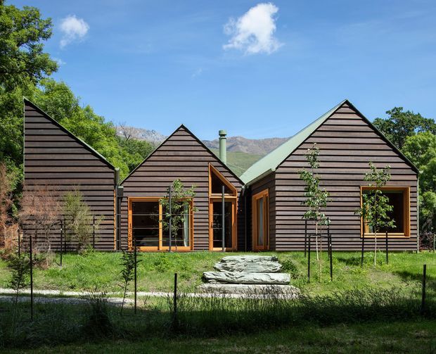 A low-rise contemporary residential building composed of several steeply gabled volumes, clad in dark horizontal timber boards with natural timber-framed glazing and pale metal roofs. The house sits within a landscaped garden, with a stone stepping path leading to the entrance and mountains visible behind it.