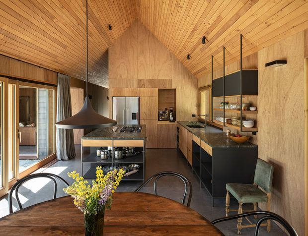 Warm, timber-lined contemporary kitchen and dining space with a dramatic pitched timber ceiling. The kitchen features dark cabinetry, stone benchtops, a central island with open shelving and cooktop, integrated stainless-steel refrigerator, and a glazed or mirrored partition displaying crockery. Full-height timber-framed glazing opens toward an exterior area, while a round dark timber dining table, black pendant light, and mixed vintage-style chairs occupy the foreground.