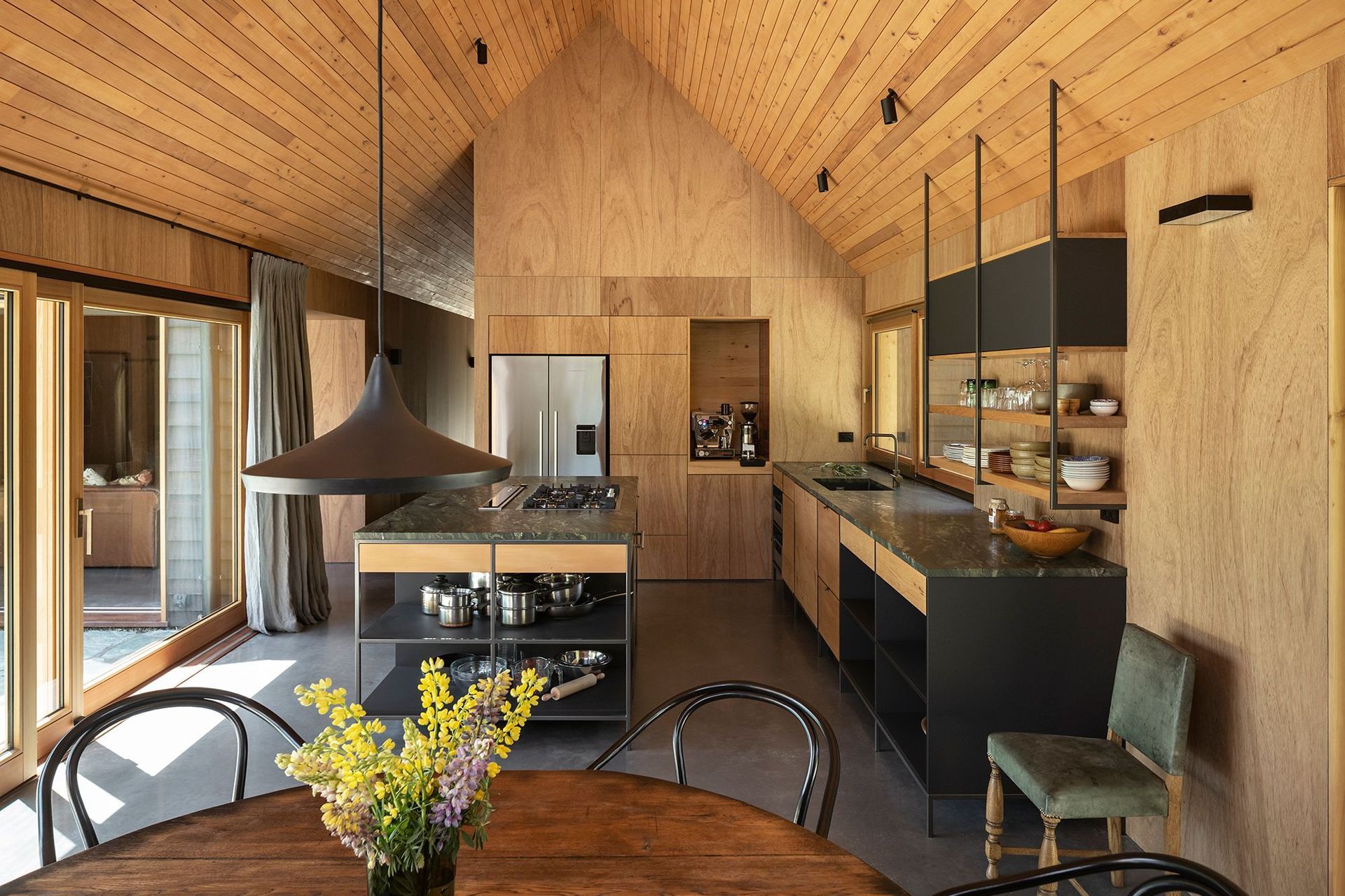 The open-plan kitchen has a wall of plywood cabinetry and an island and bench constructed in mild steel and recycled rimu, with a striking verde marble benchtop, like pounamu.