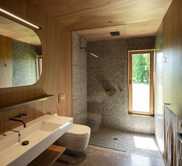 A compact, refined bathroom with a walk-in shower clad in small speckled stone-like tiles, a wall-mounted toilet, and a sculptural white basin on timber joinery. Warm timber panels cover the ceiling and walls, complemented by a rounded wall mirror, black tapware, a timber-framed window, and a heated towel rail. Natural light and greenery are visible through the window.