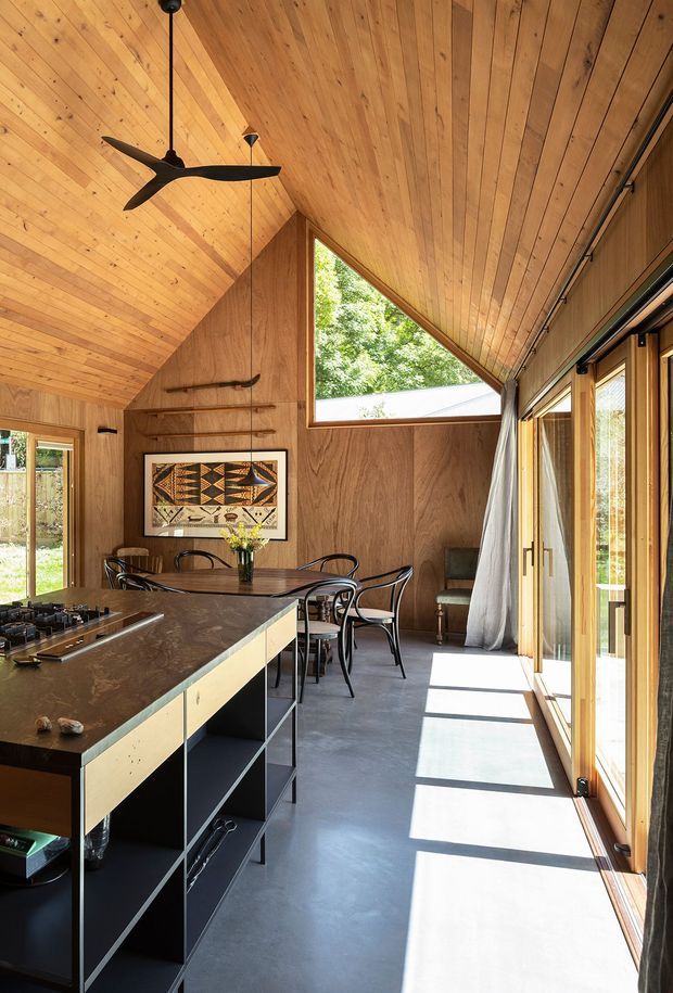 A warm, timber-lined open-plan kitchen and dining space with a dramatic sloping, vaulted ceiling. The room features a dark stone island with open shelving, a dining table and chairs, extensive full-height glazed doors, a triangular clerestory window, polished concrete flooring, and a sculptural ceiling fan. Strong sunlight enters through the glazing, creating a bright connection to the outdoors.