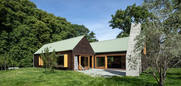 A low-rise contemporary house set within a leafy, wooded landscape. The dark horizontal timber-clad wings have steep pitched green metal roofs, large timber-framed glazed openings, and a prominent tall pale concrete chimney or masonry feature at the front. A paved entry terrace and open garage or sheltered recess are visible, with mature trees and lawn surrounding the building.