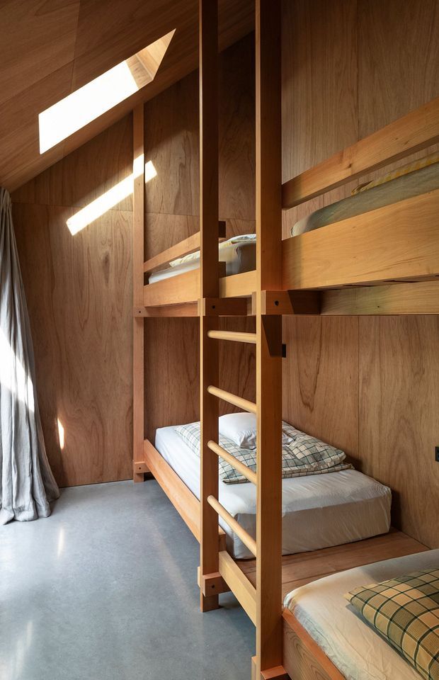 A compact bunk bedroom lined throughout with warm timber wall and ceiling panels. Built-in wooden bunk beds with integrated ladder occupy the right side, with neatly made beds, plaid bedding, and a small high-level skylight casting strong geometric sunlight across the timber surfaces. A grey polished floor and full-length curtain complete the simple, warm sleeping space.