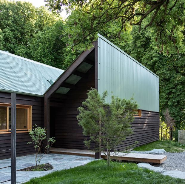 Exterior view of a compact contemporary cabin-style building set among dense mature trees. The structure combines dark horizontal timber cladding with pale corrugated metal roofing and a tall corrugated-metal gable wall. A covered entry recess, timber-framed glazing, gravel paths, stone paving, lawn, and established planting define the landscaped approach.