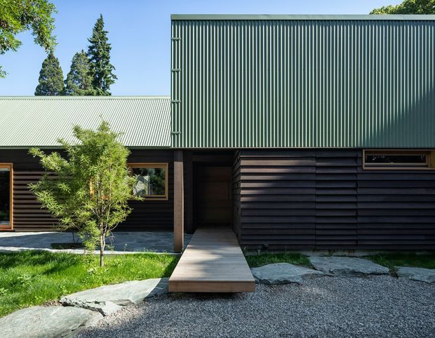 Exterior view of a low-profile contemporary building composed of dark horizontal timber cladding, a tall dark green vertically corrugated metal volume, and a pale corrugated roof. A straight timber walkway leads through a recessed entrance, with gravel landscaping, natural stone edging, lawn, and established trees in the foreground.