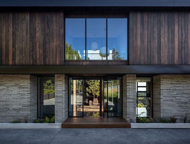 Front exterior view of a contemporary two-storey residence. The facade combines dark vertical timber cladding on the upper level with horizontal grey brickwork below, broad black-framed glazing, and a recessed glazed entry sheltered beneath a projecting upper floor. A small timber entry platform and minimalist landscaping complete the composition.
