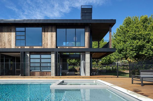 Exterior view of a contemporary two-storey residence beside a swimming pool. The home has a strong horizontal flat-roof form, dark timber cladding, expansive black-framed glazing, covered outdoor terraces, and an upper-level balcony. A timber pool deck, metal fence, mature tree, and bright blue sky complete the scene.