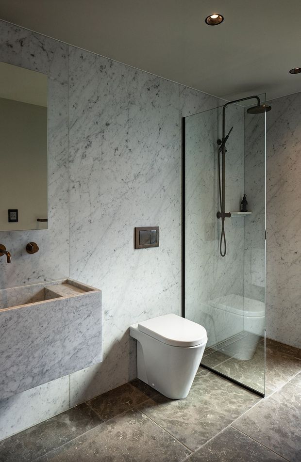 A refined contemporary bathroom featuring full-height white-grey veined marble wall cladding, dark stone-look floor tiles, a wall-hung white toilet, a floating marble vanity with integrated basin, and a frameless glass shower enclosure. Dark bronze or black fixtures, recessed ceiling spotlights, and a large wall mirror create a restrained, luxurious atmosphere.