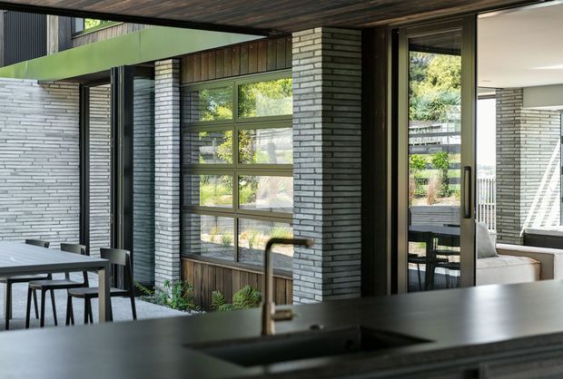 Contemporary open-plan kitchen and dining area with a dark benchtop and undermount sink in the foreground. The space opens to a covered outdoor area through large glazed doors, with exposed dark timber ceiling lining, charcoal window and door frames, grey-white horizontal brick walls, and timber accents around the glazing. A dining table and chairs sit beside the windows, while a light sofa is visible beyond the open doors.