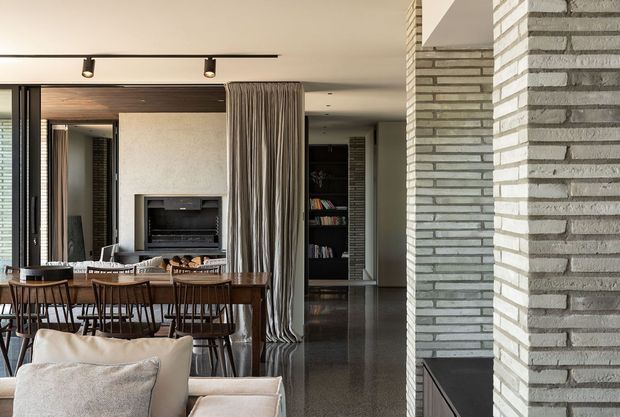 A refined open-plan living and dining area with a long timber dining table, upholstered chairs, a neutral sofa in the foreground, and a recessed fireplace set into a textured plaster wall. Full-height curtains separate the living zone from a darker hallway or library area, while a prominent pale grey brick-clad structural wall frames the right side. The space has dark polished flooring, warm timber detailing, integrated shelving, and track lighting.