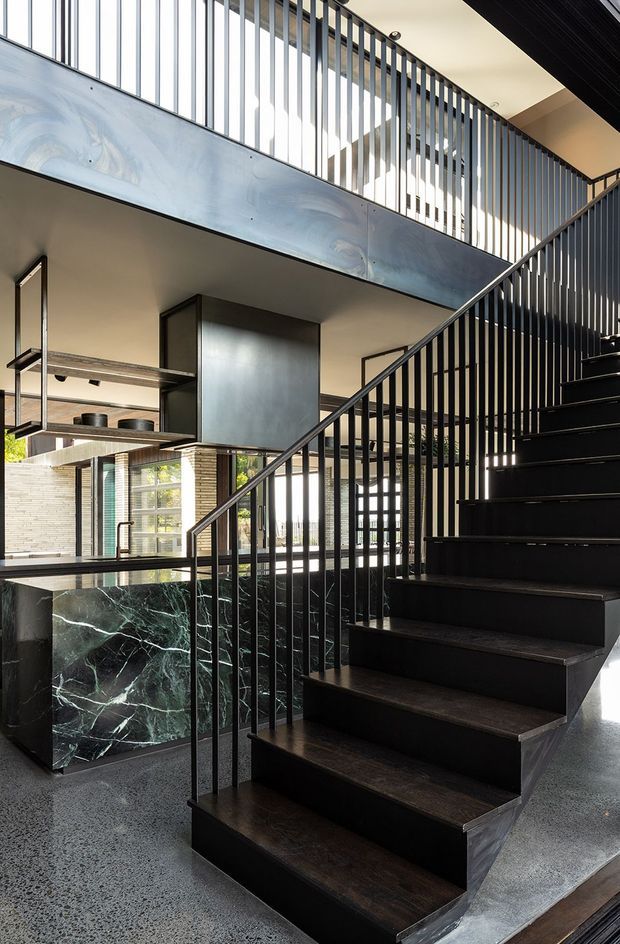 A dramatic contemporary interior centered on a dark timber staircase with slender black metal balustrades. The stair rises beside an open-plan kitchen or living area with a large veined dark stone island, exposed metal shelving, extensive glazing, and an upper-level void or mezzanine. Polished speckled flooring, pale walls, and strong natural light contrast with the dark stair and joinery.