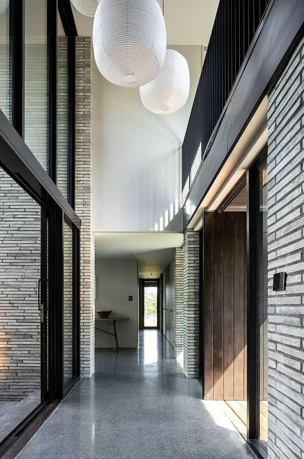 A tall, refined residential hallway or entry passage with a dramatic double-height void. The space features exposed pale grey brick walls, dark metal-framed glazing and doors, a polished grey floor, and large white ribbed pendant lights suspended overhead. Strong daylight enters from the glazing and creates rhythmic shadows across the white walls, while a timber door and compact console table add warmth. The corridor leads toward a glazed exterior opening at the far end.