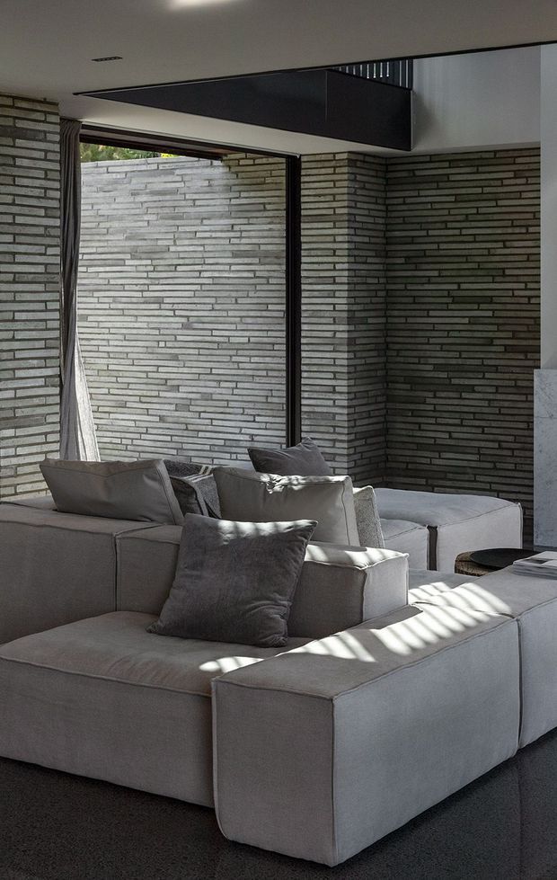 A contemporary living room with a large modular sectional sofa in pale grey upholstery, arranged around a low dark round coffee table. Exposed linear grey brick walls create a strong textured backdrop, while large glazing, curtains, and angled sunlight add contrast. A dark suspended architectural ceiling feature and black-framed openings reinforce the modern, minimalist character.