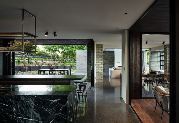A sophisticated open-plan kitchen and dining interior with a large dark marble island, black cabinetry, and bar stools in the foreground. The space opens to a landscaped outdoor courtyard through extensive glazing and also connects to a dining area with timber flooring and dark timber-framed openings. Exposed pale brick walls, polished concrete flooring, recessed and track lighting, and a restrained charcoal, grey, and timber palette create a refined contemporary atmosphere.