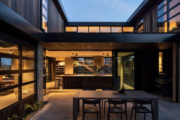 A dramatic contemporary courtyard dining and entertaining area viewed from outside, framed by dark timber-clad upper levels and extensive black-framed glazing. The covered ground-floor space contains a large outdoor dining table, an illuminated indoor kitchen and bar visible through the opening, and warm integrated lighting. The composition emphasizes a sheltered courtyard, strong geometric lines, and a central open-to-sky void.