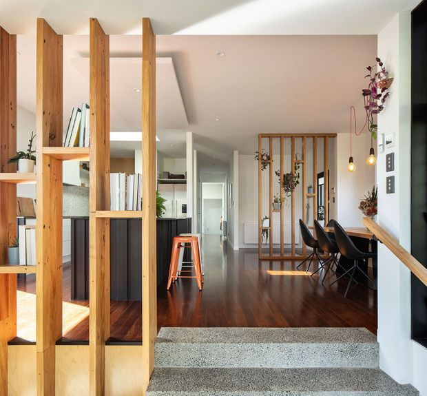 A bright, open-plan contemporary interior viewed from a carpeted entry level, with two concrete-look steps leading into a timber-floored living and dining zone. Vertical natural timber screens and shelving divide the entry from the kitchen and dining areas. The kitchen has dark fixed cabinetry and a pale benchtop, while the dining area features a timber table, black chairs, and hanging pendant lights. White walls, recessed ceiling details, indoor plants, and an open hallway create a warm, airy atmosphere.