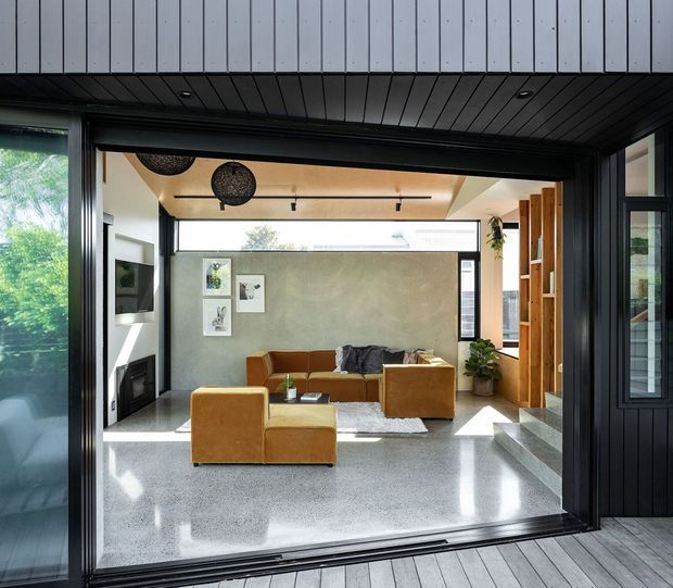 A contemporary living room viewed from an exterior deck through a large open glazed opening. The room features a polished terrazzo floor, warm tan modular sofas and armchairs, a textured grey feature wall, floor-to-ceiling glazing, black-framed doors and windows, timber shelving, and a recessed ceiling with track lighting. The dark vertical exterior cladding and generous opening create a strong indoor-outdoor connection.