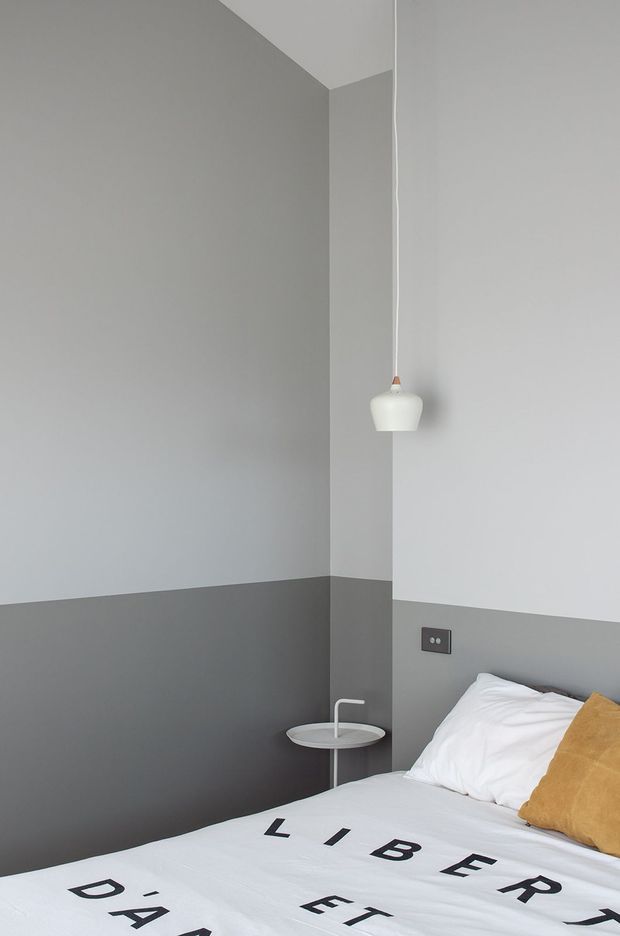 Minimalist bedroom corner with a white bed dressed in graphic lettering, a mustard accent cushion, two-tone grey wall treatment, a small round bedside table, and a simple white pendant light. The room has clean lines, sparse furnishings, and a cool neutral palette with a warm mustard accent.