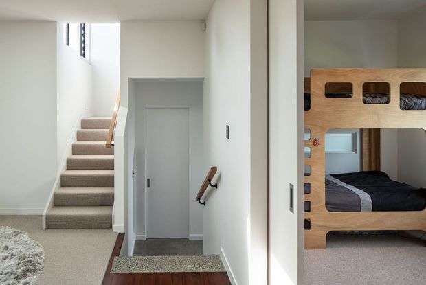 Bright, minimalist residential hallway and stair landing with white walls, carpeted stairs and floors, simple timber handrails, and a closed interior door. A timber bunk bed with integrated storage and a bed is visible through the opening on the right, indicating an adjacent bedroom or kids' room.