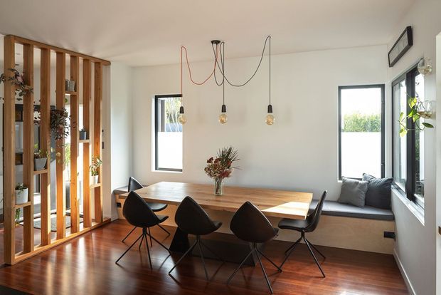 Bright contemporary dining area with a long timber dining table and four dark sculptural chairs, set beside a built-in window seat. Warm timber flooring and a vertical timber-screen partition with integrated shelving and plants define the room. White walls, black-framed windows, and exposed pendant cords with individual bulbs create a clean, relaxed interior.