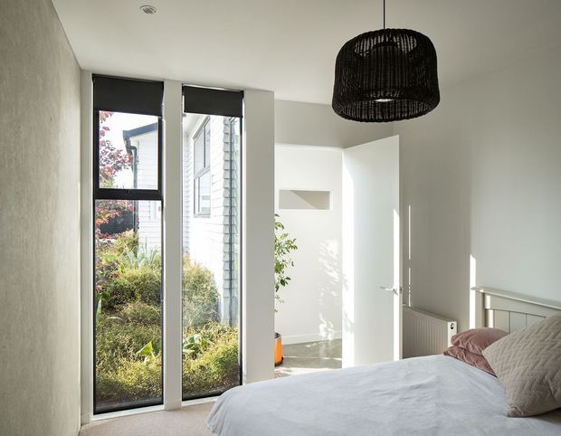 Bright contemporary bedroom with a neatly made bed, white walls and ceiling, tall narrow black-framed windows with roller blinds, and a large black woven pendant light. A white door opens to an adjacent area, while a textured neutral wall and radiator add subtle detail. Garden planting and neighbouring cladding are visible outside the windows.