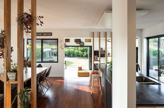Open-plan contemporary interior combining a kitchen, dining area and living space. The room features dark timber flooring, white walls and ceiling, warm exposed timber divider posts with integrated shelving and plants, a dark kitchen island with stools, and large windows and glazed doors opening to a landscaped outdoor area. Natural light creates a warm, inviting atmosphere.