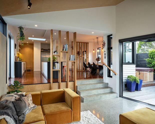 A bright, open-plan contemporary home interior viewed from the living room, with a tan sectional sofa in the foreground, polished concrete steps and flooring, timber-lined ceiling, and a timber open shelving and screen structure dividing the living, kitchen, and dining zones. Large black-framed glazing opens toward an outdoor deck with planters and a spa. The space features warm timber, neutral finishes, indoor plants, and layered ceiling and pendant lighting.