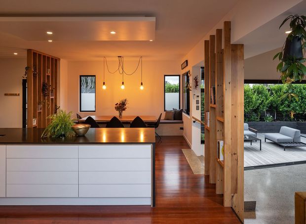 Open-plan contemporary kitchen and dining area viewed from the kitchen island, with white handleless cabinetry, a dark benchtop, warm timber flooring, a timber slatted divider and built-in shelving. A dining table, pendant lights and living seating are visible, with large openings leading to a paved outdoor terrace and landscaped garden.