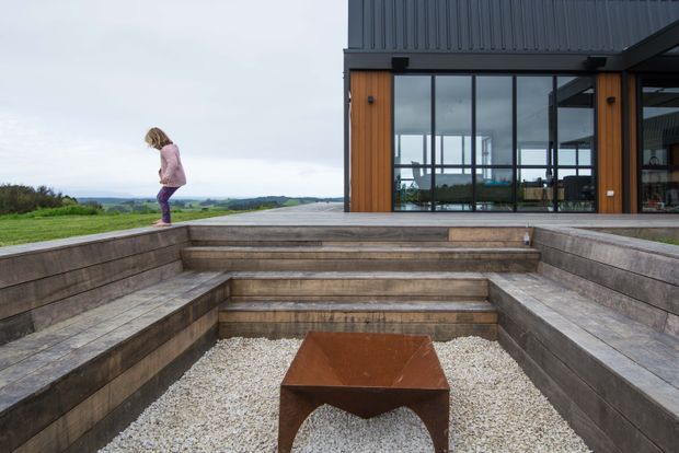 A contemporary dark-clad house opens onto a broad stepped outdoor terrace and sunken gravel courtyard. Timber-lined walls, expansive black-framed glazing, built-in timber seating, and a rusted steel fire-pit table create a structured outdoor entertaining area, with open rural landscape beyond.