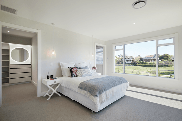 Bright, contemporary bedroom with a low-profile upholstered bed, layered white and grey bedding, a small white cross-leg bedside table, and a built-in wardrobe visible through the open dressing area. The room has light neutral finishes, recessed lighting, pendant bedside lights, broad windows, and views toward a landscaped suburban setting.