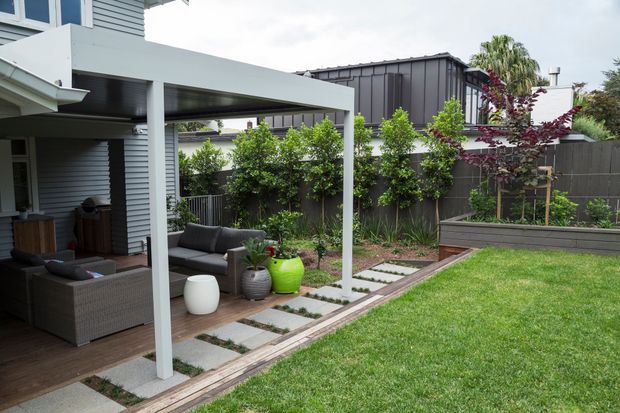A contemporary landscaped backyard with a covered outdoor patio adjoining a modern home. The patio has dark woven outdoor seating, timber decking, a white structural pergola frame, and a paved stepping-stone path leading through a lawn and planted garden beds. Grey horizontal cladding, dark fencing, raised planters, and sculptural outdoor pots complete the restrained garden design.