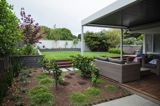 A landscaped backyard with a covered outdoor lounge area adjoining the house. The space includes a timber deck, woven outdoor seating, a large flat-roofed canopy supported by slim posts, raised garden beds, lawn, shrubs, and a concrete-paved path.