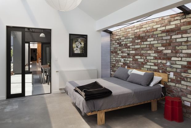 A contemporary bedroom with a low timber bed dressed in grey bedding, set against a striking exposed reclaimed-brick feature wall. White walls, a concrete-look floor, black-framed glazed doors, a radiator, and a simple pendant light create an understated industrial character. A dining or living area is visible through the internal glazing.