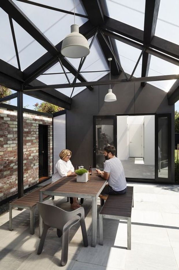 A bright, contemporary enclosed outdoor dining area or courtyard with a dramatic glazed, exposed steel-framed roof. The space has a dark painted structure, translucent roof panels, pendant lights, exposed brick walls, concrete paving, and black-framed glazed doors opening toward a white interior. A timber dining table with built-in bench seating and chairs occupies the foreground.
