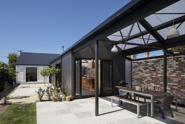 A contemporary single-storey home extension with a covered outdoor dining terrace. The dark metal-framed structure has an open steel-truss roof, large glazed doors and windows, and pendant lights over a long outdoor dining table. A restored-looking exposed brick wall forms the rear boundary, while pale paved flooring, planted garden beds, and a small adjoining gabled section are visible.