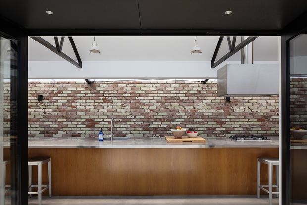 A contemporary kitchen viewed through wide black-framed glazing or doors. The space features a long timber island with a pale stone benchtop, integrated sink and cooktop, two white stools, an exposed red-and-white brick splashback wall, dark ceiling panels, and exposed dark structural beams beneath a high ceiling. Pendant lights and a large white rangehood are visible.