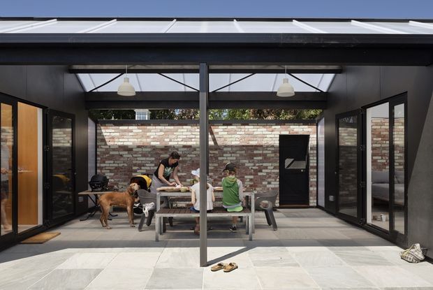 A covered outdoor entertaining area enclosed by dark-framed glazed doors, with a translucent pitched roof, exposed central support post, brick feature wall, paved floor, dining table and benches, and people and dogs gathered around the table. The space connects directly to interior rooms on both sides and has a relaxed indoor-outdoor character.