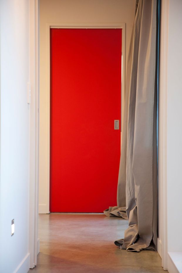 A narrow, minimalist interior hallway viewed toward a vivid red flush door. The corridor has smooth pale walls, white door trim, a polished warm-toned concrete floor, and tall loose grey-beige curtains hanging along the right side. A small wall switch or outlet is visible on the left, with soft natural light entering from the side.