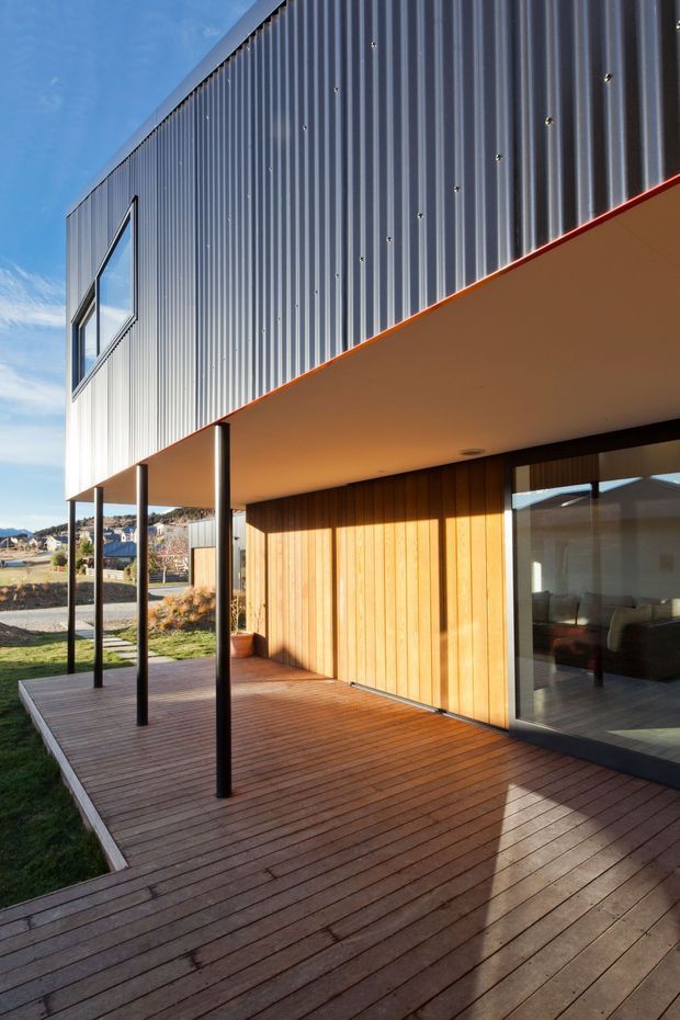 Exterior view of a contemporary two-storey house with a corrugated metal upper facade, warm vertical timber wall cladding, slim dark window and door frames, and a broad timber deck beneath a projecting upper level. The covered deck is supported by slender black posts and opens toward a landscaped suburban setting.