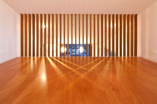 A warm, contemporary interior with a polished timber floor and ceiling, framed by white walls. A full-width vertical timber slat screen forms a balustrade or partition overlooking a lower-level area, with integrated lighting creating strong linear shadows across the floor.