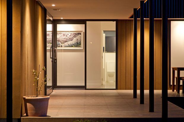 A warmly lit contemporary interior hallway or entry passage with timber wall panelling, a broad timber floor, black-framed glazed doors, and a dark vertical slatted partition. A framed artwork and potted plant add restrained decoration, while a bathroom is visible through the glazing and a dining area is partially visible at right.
