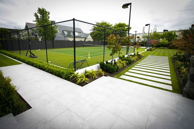 Contemporary landscaped outdoor area with a fenced synthetic-turf tennis court, broad pale tiled paving, and a linear stepping-stone garden path bordered by clipped hedges and ornamental planting. Modern black lighting poles and privacy fencing frame the recreational space.