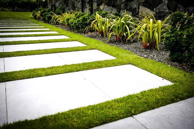 A landscaped garden walkway composed of large rectangular white concrete stepping slabs set into neatly trimmed grass. The path runs alongside a dark natural-stone retaining wall with layered ornamental grasses and dense green planting, creating a clean, contemporary outdoor approach.