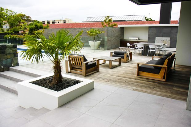 Contemporary outdoor entertaining terrace with a timber-decked lounge area, wood-framed black-cushioned armchairs, coffee tables and an outdoor dining setting. Large pale porcelain-tiled paving surrounds a raised white planter containing a palm, while dark grey stone-clad walls and a low red-tiled roof define the backdrop. Steps lead toward a partially visible pool area.